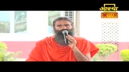 A man in an orange robe holds a microphone, speaking into it. The background suggests a broadcast from India, possibly on Aastha Bhajan.
