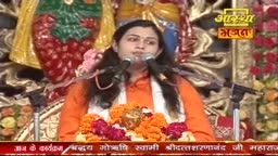 A woman in saffron, with a white scarf, speaks into two microphones, likely on Aastha Bhajan. Below, a garland of orange and red flowers rests on the table in front of her.
