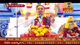 A man in India, wearing garlands and a shawl, is speaking into a microphone, likely leading an Aastha Bhajan. Brightly colored flowers and decorations surround him.
