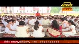 A large group of people, mostly in white, sit on the ground, facing a man in a light-colored outfit. The scene is likely part of an Aastha Bhajan broadcast from India.
