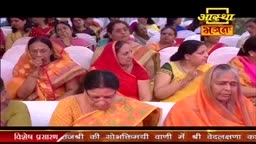 Women in vibrant saris sit in white chairs, some with hands clasped, others with fingers pressed to their lips. The Aastha Bhajan logo is visible above them, and a red banner with Hindi text runs along the bottom of the screen.

