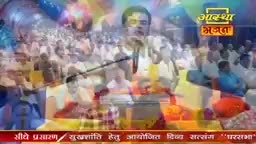 A man in a white shirt holds a microphone, speaking to a large crowd. The Aastha Bhajan logo sits in the corner, and the scene seems to be a live broadcast from India.
