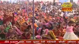 A massive crowd, mostly women in vibrant saris, is gathered, many with hands raised. A little girl in a pink dress dances near the front, while the Aastha Bhajan logo is visible in the background.
