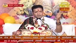 A man in a white shirt, with a red mark on his forehead, is speaking into a microphone. He gestures with his right hand, while a garland of flowers rests around his neck.
