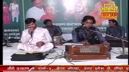 Two men sit cross-legged, singing into microphones, while another plays a harmonium. The backdrop shows a banner for Aastha Bhajan, a religious program from India.
