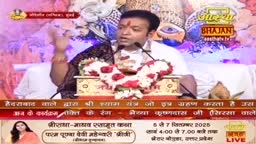 The man at the podium gestures emphatically, his hand raised as he speaks into two microphones. A garland of flowers and a small wooden box sit on the table in front of him, with the Aastha Bhajan logo visible above.
