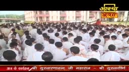 A large crowd in white clothing sits on the ground, facing a speaker. The Aastha Bhajan logo sits in the top right corner, and a banner with Hindi text runs across the bottom of the screen.
