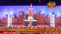 A man in a white shirt sits at a desk, speaking in front of a large statue of a deity. The stage is set for a broadcast, with the "Aastha Bhajan" logo visible above.
