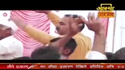 A man in a yellow shirt raises his arms, his hands open, as if in prayer or praise. The Aastha Bhajan logo is visible in the upper right corner.
