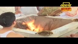 Flames leap from a square pit as people sit around it, some holding small plates. The scene is part of an Aastha Bhajan broadcast in India.

