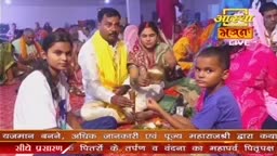 A family sits together, a small boy holding something out toward the camera. The scene is part of what looks like a live broadcast, with the "Aastha Bhajan" logo visible in the corner.

