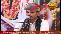 A man in a vibrant turban sings into a microphone, likely for an Indian devotional program. The Aastha Bhajan logo is visible, and a garland of flowers hangs in the background.
