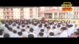 A large group of people in white clothing are seated on the ground, facing a person in red. The Aastha Bhajan logo is visible at the top of the frame.
