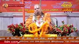 A man in saffron robes sits at a table, speaking into a microphone. He's surrounded by floral arrangements and a garland, with an "Aastha Bhajan" banner visible in the background.
