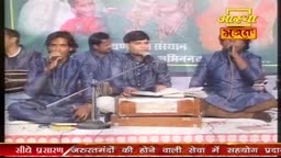 Men in blue shirts sit cross-legged, singing into microphones during a live Aastha Bhajan broadcast. A harmonium sits open in front of one of the performers.
