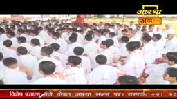 A large group of people, all dressed in white, sit close together on the ground. The Aastha Bhajan logo is visible at the top, indicating a religious gathering in India.
