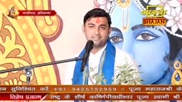 A man with a red mark on his forehead stands at a microphone, speaking on the Aastha Bhajan channel. Behind him, a large image of Krishna is visible, and a small plant sits to his right.
