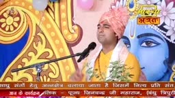 A man in a turban and yellow shirt speaks into a microphone, likely during an Aastha Bhajan event in India. Behind him, a large blue image of a deity provides a backdrop for the broadcast.
