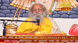 A man with a long white beard and glasses sits behind a desk, speaking into a microphone. The Aastha Bhajan logo is visible, and a banner with contact information is displayed at the bottom.
