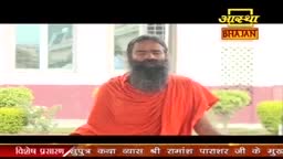 A man in an orange robe sits before a camera, his long, dark beard prominent against the backdrop of a building. The Aastha Bhajan logo is visible at the top of the screen.
