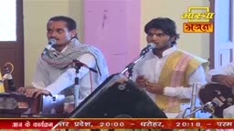 Two men, dressed in white, sing into microphones, one with a scarf draped around his neck. The Aastha Bhajan logo glows above them as they perform.
