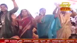 Women in colorful saris are clapping and raising their arms, seemingly enjoying a performance. The logo for Aastha Bhajan is visible, suggesting this is a religious event in India.
