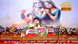 A man in a yellow shirt speaks into a microphone, framed by a floral arrangement. Behind him, a large screen displays a painting of Krishna and Radha, with the Aastha Bhajan logo in the corner.
