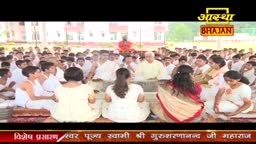 A large group of people, many in white, sit on the ground, facing a seated leader. The Aastha Bhajan logo is visible at the top of the screen.
