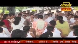 A large group of people are gathered, many wearing white, in what looks like a public space.  The Aastha Bhajan logo is visible, suggesting a religious event is underway in India.
