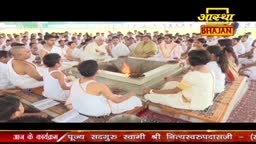 Around a central fire, a large group of people in white clothing are seated, facing inward. The Aastha Bhajan logo is visible above the gathering.
