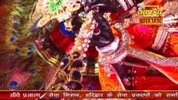 A dark-skinned figure, adorned with jewels, is holding a garland, likely as part of a devotional ceremony. Peacock feathers and vibrant red fabrics frame the scene, hinting at a celebration within India, possibly a broadcast by Aastha Bhajan.
