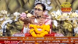 A woman in glasses and a headpiece speaks into a microphone, her hands gesturing as she addresses the audience. A bright yellow garland and the Aastha Bhajan logo are in the foreground, with text in Hindi at the bottom of the screen.

