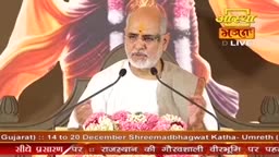 A man with a white beard and a tilak on his forehead speaks into two microphones, gesturing with his hands. Behind him, a backdrop of flowing fabrics and the Aastha Bhajan logo creates a colorful stage for the event.
