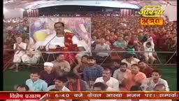 A man in a white kurta speaks into a microphone as a massive crowd watches, many with their hands clasped in prayer. The event is broadcast on the Aastha Bhajan channel, with a banner in Hindi at the bottom of the screen.
