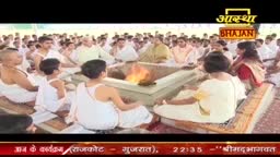 People in white clothes sit in a circle around a small fire, likely part of a religious ceremony. The Aastha Bhajan logo is visible in the upper right corner, indicating this is a broadcast from India.
