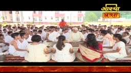 A large group of people in white clothing are seated on the ground, listening to a man in a light-colored shirt. The scene appears to be part of an Aastha Bhajan gathering in India, with the channel's logo visible in the top right.
