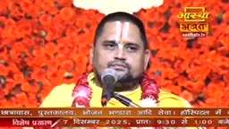 A man with a prominent tilak speaks into a microphone, a garland draped around his neck. Behind him, a wall of bright orange flowers provides a vibrant backdrop for the Aastha Bhajan broadcast.
