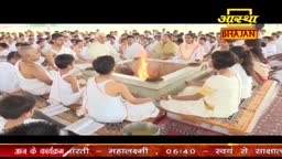 People in white clothing sit on the floor, facing a small fire pit at the center of the gathering. The Aastha Bhajan logo is visible at the top, and the scene appears to be part of a religious ceremony in India.
