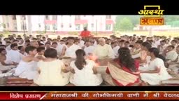 People dressed in white sit in a large circle, many facing a small fire. Above them, the Aastha Bhajan logo is visible, indicating a broadcast from India.

