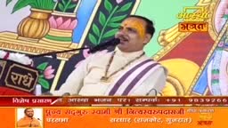 A man in a cream-colored outfit, with a yellow mark on his forehead, speaks into a microphone. Behind him, colorful artwork and the logo for Aastha Bhajan are visible.
