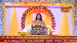 A man in a beige robe gestures from behind a lectern, speaking into a microphone. The backdrop, framed by flowers and draped with yellow fabric, features the logo for Aastha Bhajan, a devotional program from India.
