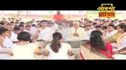 A large group of people are gathered, many in white, facing forward. In the background, a man in orange sits elevated, while a man in a yellow shirt stands near the front.
