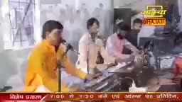 A man in a saffron-colored shirt sings into a microphone, while others play instruments in the background. The scene, likely a broadcast from India, features a musical performance on Aastha Bhajan.
