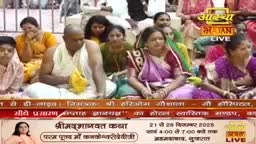 People are seated on the floor, listening intently, some with their hands clasped. The Aastha Bhajan logo is visible, indicating a live broadcast of a religious event in India.
