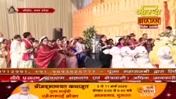 A group of people in traditional Indian attire are gathered, some holding microphones. A woman in a red sari stands prominently, facing the crowd.