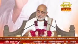 A man with a shaved head, wearing a white kurta and a garland of red flowers, speaks into a microphone. A banner with Hindi text and the Aastha Bhajan logo is visible behind him.