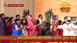 A group of people are gathered, some holding plates with offerings. A woman in a bright pink sari is in the center, her hands extended.