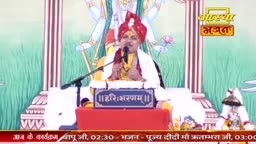 A man in a red turban speaks into a microphone, his hands gesturing. Behind him, a large, stylized image of a deity stands against a backdrop of green and white stripes.