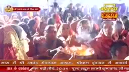 A crowd of people, many in red and saffron, gather around a central fire, with smoke rising. This is part of a devotional gathering, perhaps a Bhajan, in India.