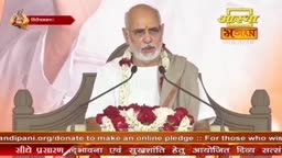 A man in white speaks into two microphones, his head adorned with a garland of white and red flowers. Behind him, a banner displays the Aastha Bhajan logo.